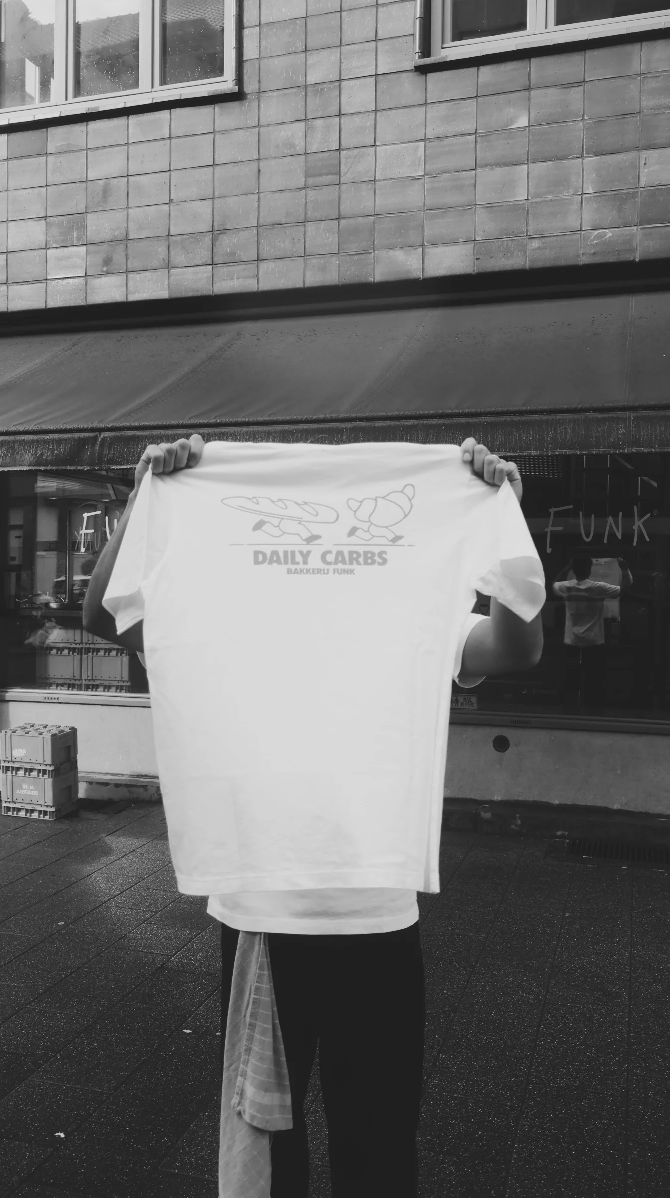 A person holding up a 'Daily Carbs' Bakkerij FUNK t-shirt outside the bakery